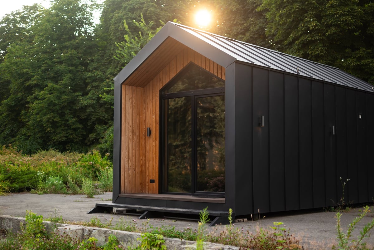 Modern tiny house with wooden and black metal facade, surrounded by greenery, lit by soft evening sunlight